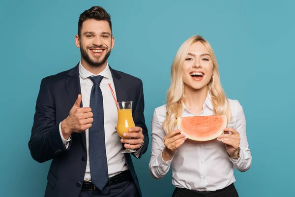 Feliz hombre de negocios con jugo de naranja mostrando golpe hasta cerca sonriente mujer de negocios con jugosa sandía sobre fondo azul - foto de stock