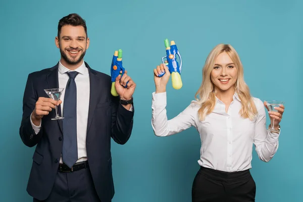 Empresarios felices sosteniendo vasos de cóctel y pistolas de agua de juguete sobre fondo azul - foto de stock