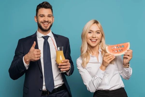 Felice uomo d'affari con succo d'arancia mostrando pollice verso l'alto vicino donna d'affari sorridente con ciao iscrizione estiva su fetta di anguria su sfondo blu — Foto stock