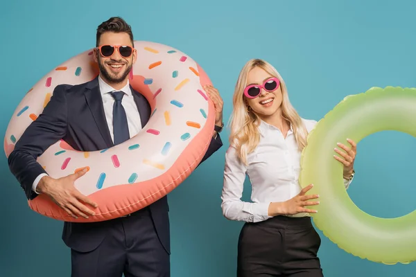 Glückliche Geschäftsleute mit Sonnenbrille, die aufblasbare Ringe halten, während sie in die Kamera auf blauem Hintergrund schauen — Stockfoto