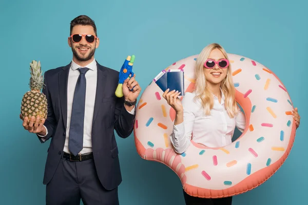 Счастливые бизнесмены с плавательным кольцом, документами, водяным пистолетом и свежим ананасом, улыбающиеся в камеру на синем фоне — стоковое фото