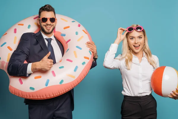 Homme d'affaires heureux dans l'anneau de natation montrant pouce vers le haut près souriant femme d'affaires tenant ballon gonflable sur fond bleu — Photo de stock