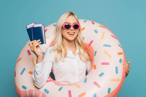 Happy businesswoman in sunglasses and swim ring holding passports and air tickets isolated on blue — Stock Photo