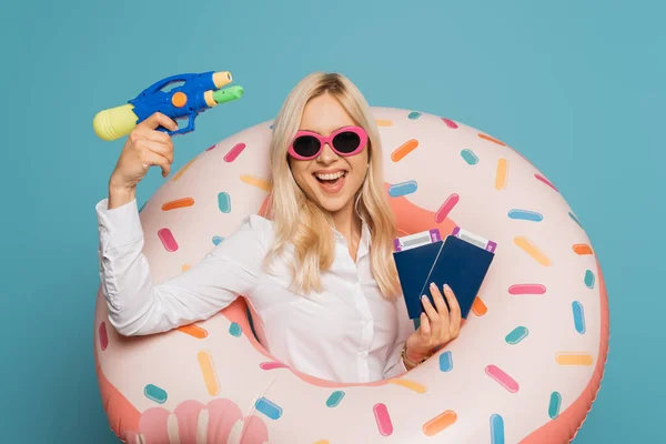 Cheerful businesswoman in sunglasses and swim ring holding passports, air tickets and water gun isolated on blue — Stock Photo