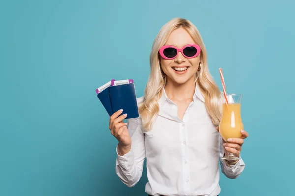 Feliz mujer de negocios en gafas de sol sosteniendo vaso de jugo de naranja, pasaportes y billetes de avión sobre fondo azul - foto de stock