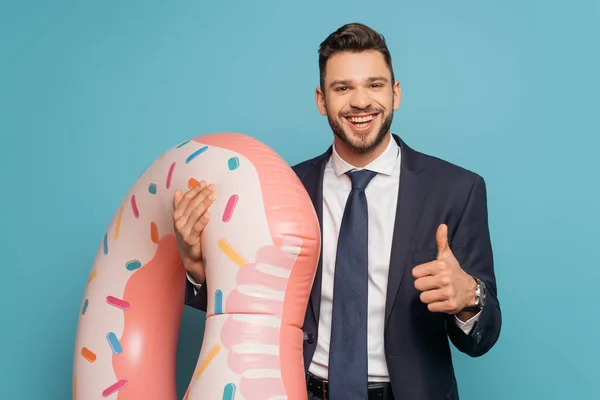 Feliz hombre de negocios mostrando el pulgar hacia arriba mientras sostiene el anillo inflable sobre fondo azul - foto de stock