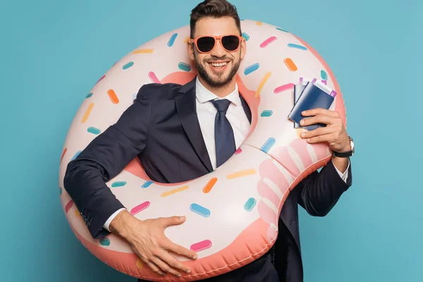 Joyful businessman in swim ring holding passport and ait tickets on blue background — Stock Photo