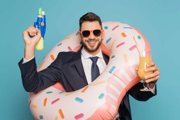 Alegre hombre de negocios en anillo de natación sosteniendo pistola de agua y vaso de jugo de naranja sobre fondo azul - foto de stock