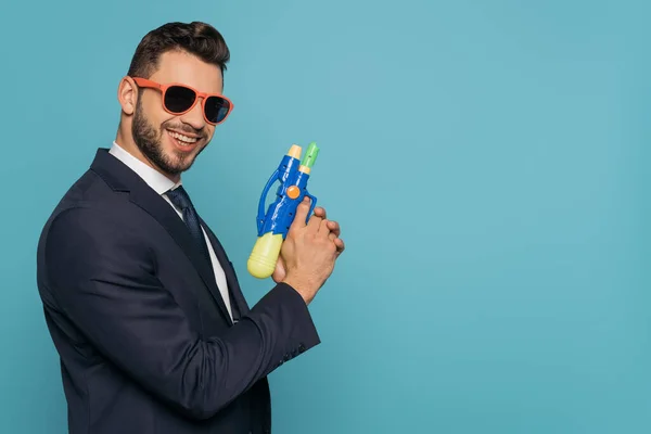 Freudiger Geschäftsmann hält Wasserpistole in der Hand, während er isoliert auf blau in die Kamera blickt — Stockfoto