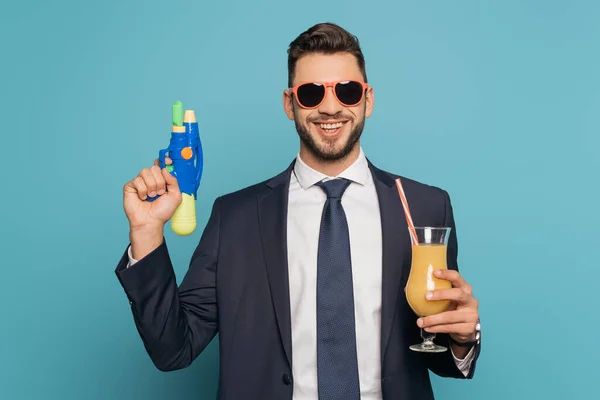 Feliz hombre de negocios sosteniendo vaso de cóctel con jugo de naranja y pistola de agua sobre fondo azul - foto de stock