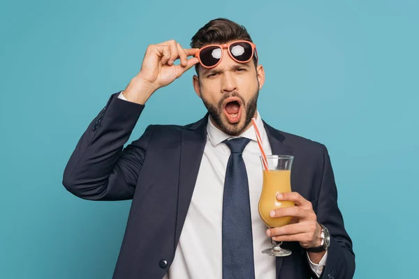 Sorprendido hombre de negocios con la boca abierta tocando gafas de sol mientras sostiene el jugo de naranja sobre fondo azul - foto de stock