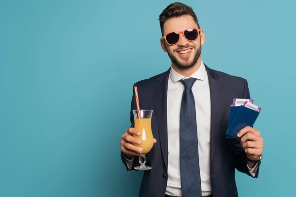 Hombre de negocios sonriente con gafas de sol con pasaportes, billetes de avión y zumo de naranja sobre fondo azul - foto de stock