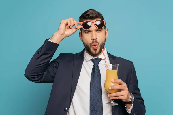 Homme d'affaires surpris touchant lunettes de soleil tout en tenant verre à cocktail avec du jus d'orange isolé sur bleu — Photo de stock