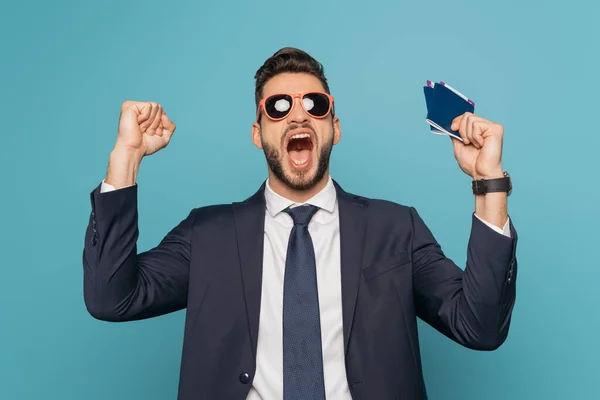 Emocionado hombre de negocios en gafas de sol gritando y mostrando gesto ganador mientras sostiene pasaportes y billetes de avión aislados en azul - foto de stock