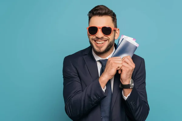 Excited businessman in sunglasses holding passports and air tickets while smiling at camera isolated on blue — Stock Photo