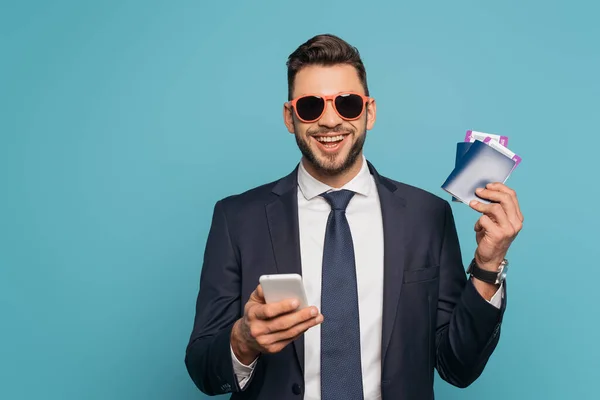 Happy businessman in sunglasses using smartphone and smiling at camera while holding passports and air tickets isolated on blue — Stock Photo