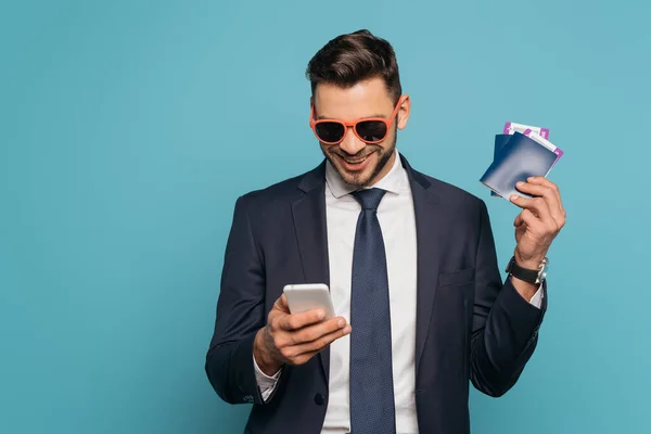 Alegre hombre de negocios en gafas de sol charlando en el teléfono inteligente, mientras que la celebración de pasaportes y billetes de avión aislados en azul - foto de stock