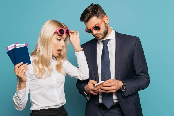 Schockierte Geschäftsfrau hält Dokumente in der Hand und berührt Sonnenbrille, während sie das Smartphone in den Händen eines Geschäftsmannes auf blauem Hintergrund betrachtet — Stockfoto