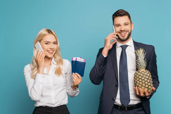 Cheerful businesspeople talking on smartphones while holding passports, air tickets and fresh pineapple on blue background — Stock Photo