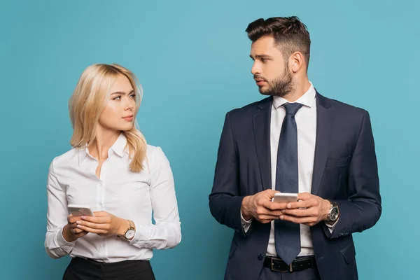 Молода бізнес-леді і бізнесмен дивиться один на одного, тримаючи смартфони на синьому фоні — стокове фото