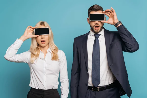 Überraschte Geschäftsfrau und Geschäftsfrau bedeckt Augen mit Smartphones mit leerem Bildschirm auf blauem Hintergrund — Stockfoto