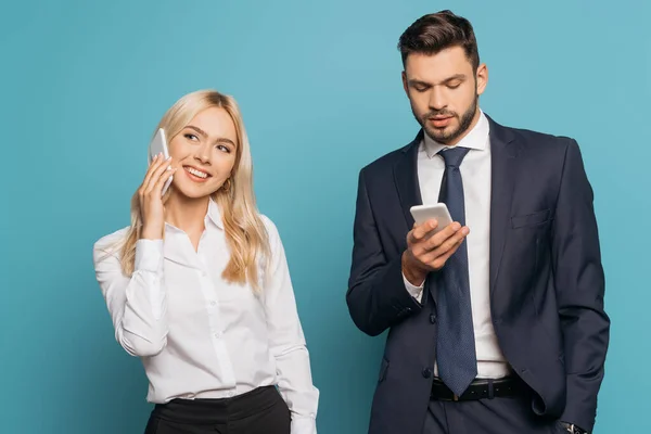 Sonriente mujer de negocios hablando en el teléfono inteligente cerca serio hombre de negocios charlando sobre fondo azul - foto de stock