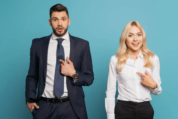 Surprised businessman and businesswoman pointing with fingers at themselves on blue background — Stock Photo
