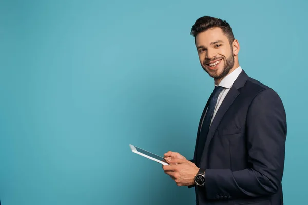 Красивый бизнесмен, использующий цифровой планшет, улыбаясь в камеру на синем фоне — стоковое фото