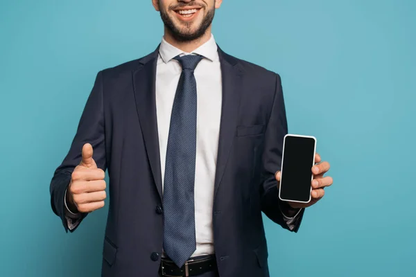 Abgeschnittene Ansicht eines lächelnden Geschäftsmannes, der den Daumen nach oben zeigt, während er sein Smartphone mit leerem Bildschirm auf blauem Hintergrund hält — Stockfoto