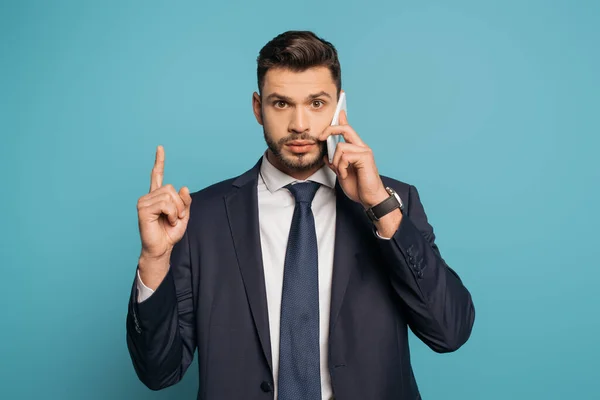 Hombre de negocios confiado hablando en el teléfono inteligente, mostrando gesto de idea y mirando a la cámara en el fondo azul - foto de stock