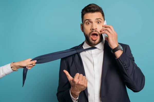 Cropped view of woman pulling tie of shocked businessman talking on smartphone and pointing with thumb on blue background — Stock Photo