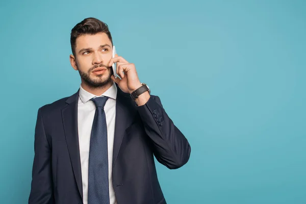 Serio, hombre de negocios guapo hablando en el teléfono inteligente y mirando hacia otro lado aislado en azul — Stock Photo