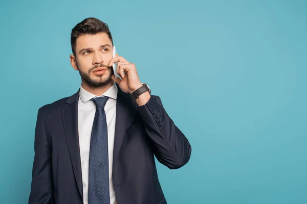 Serio, hombre de negocios guapo hablando en el teléfono inteligente y mirando hacia otro lado aislado en azul - foto de stock