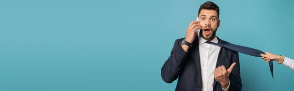 Cropped view of woman pulling tie of shocked businessman talking on smartphone and pointing with thumb on blue background, panoramic orientation — Stock Photo