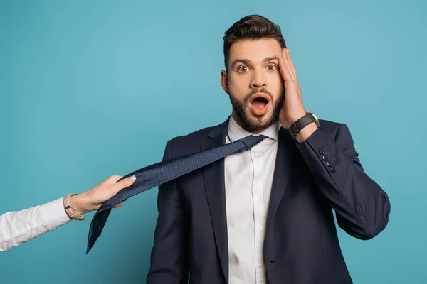 Abgeschnittene Ansicht einer Frau, die Krawatte eines schockierten Geschäftsmannes zieht, der Gesicht auf blauem Hintergrund berührt — Stockfoto