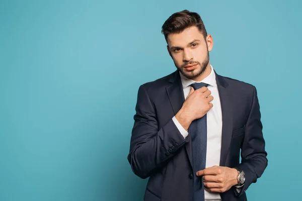 Selbstbewusster, gutaussehender Geschäftsmann, der Krawatte berührt, während er in die Kamera auf blauem Hintergrund blickt — Stockfoto