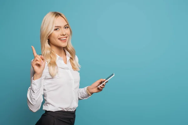 Feliz mujer de negocios mostrando gesto de idea mientras sostiene el teléfono inteligente y mirando a la cámara aislada en azul - foto de stock