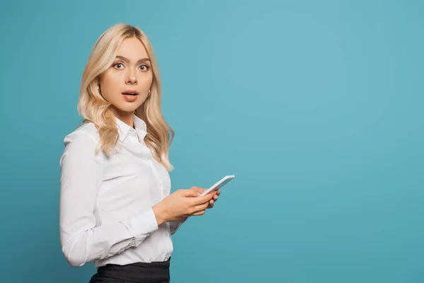 Шокированная блондинка-бизнесмен смотрит в камеру, используя смартфон, изолированный на синий — стоковое фото