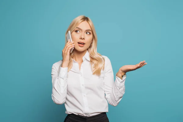 Pensativa mujer de negocios de pie con el brazo abierto mientras habla en el teléfono inteligente aislado en azul - foto de stock