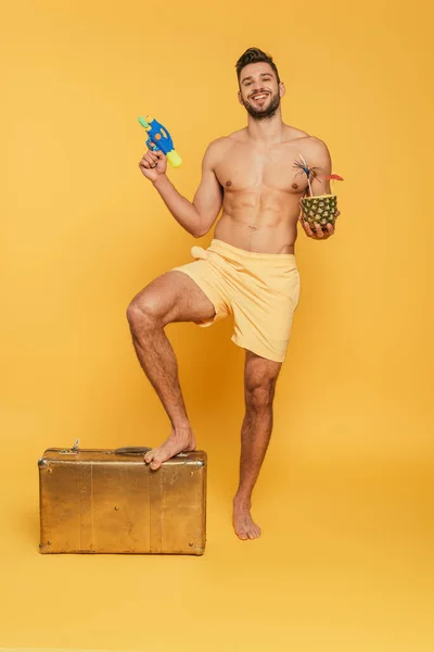 Felice uomo senza maglietta in possesso di acqua pistola cocktail in ananas mentre calpestando valigia vintage su sfondo giallo — Foto stock