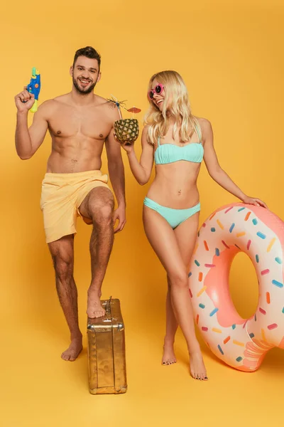 Chica feliz con anillo inflable y piña con cóctel, y hombre guapo con pistola de agua pisando la maleta vintage sobre fondo amarillo - foto de stock