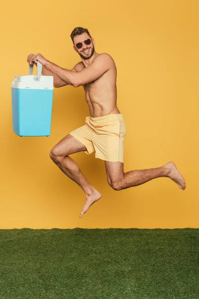 Homem sem camisa sorridente segurando geladeira portátil enquanto levita no fundo amarelo — Fotografia de Stock