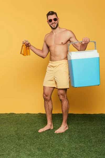 Homem sem camisa alegre segurando geladeira portátil e garrafas de cerveja no fundo amarelo — Fotografia de Stock