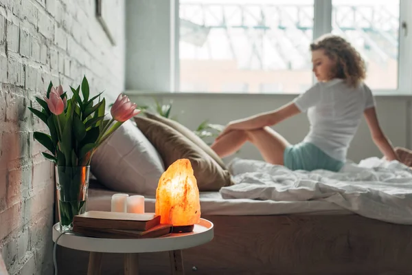 Hermosa chica en la cama con lámpara de sal del Himalaya, flores y libros en la mesa en el dormitorio, enfoque selectivo - foto de stock