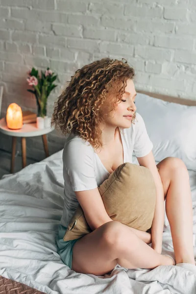 Ніжна дівчина вдома обнімає подушку на ліжку в затишній спальні з гімалайською соляною лампою. — стокове фото