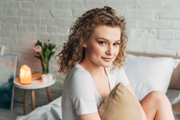 Attractive curly woman in homewear hugging pillow on bed in cozy bedroom with Himalayan salt lamp — Stock Photo