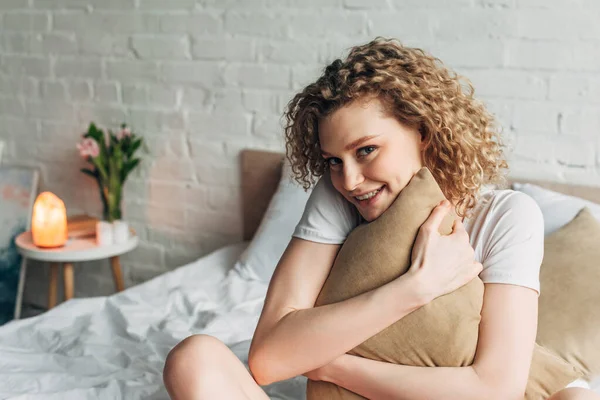 Alegre hermosa chica en ropa de casa abrazando almohada en la cama en el acogedor dormitorio con la lámpara de sal del Himalaya - foto de stock