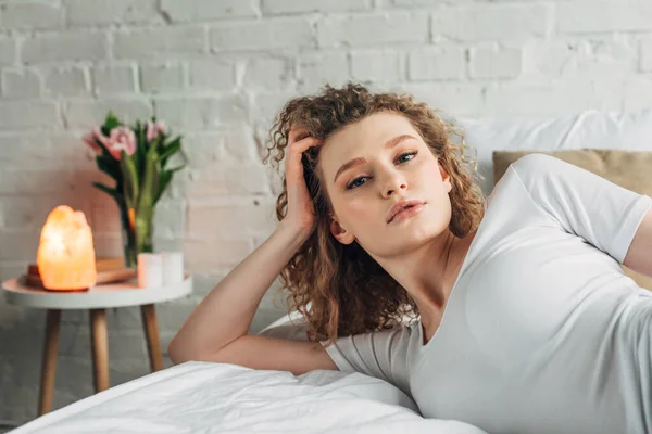 Jolie fille bouclée en vêtements de maison couché sur le lit dans la chambre à coucher avec lampe de sel himalayen — Photo de stock