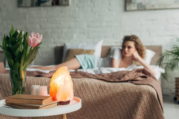 Селективное внимание девушки, отдыхающей в спальне с гималайской соляной лампой, цветами и книгами — стоковое фото
