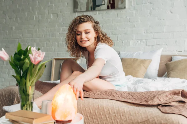 Happy woman in bedroom with Himalayan salt lamp and flowers, selective focus — Stock Photo